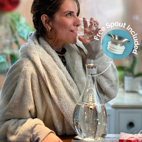 A woman in a cozy robe drinks water from a glass as the MAYU Swirl Carafe | Onyx by Mayu Water, filled with water, sits in front of her. An inset highlights the carafe’s spout with the text Free Spout Included.
