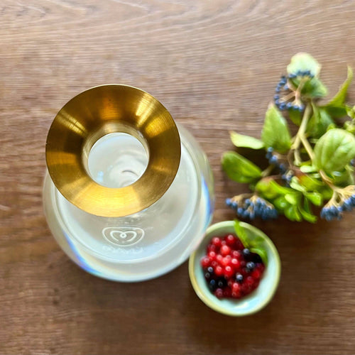 A top-down view of Mayu Water's Swirl Golden Pouring Spout on a glass bottle, alongside a bowl of red and black berries and green leaves with blue berries, all beautifully arranged on a wooden surface.