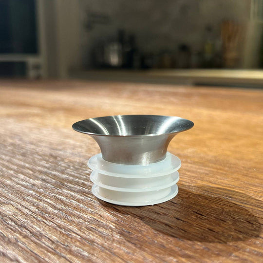 A close-up of the Mayu Water Swirl Stainless Steel Pouring Spout with its wide, flared top rests on a wooden surface, ideal for pairing with a glass carafe in modern table decor. A blurred kitchen background adds a stylish accent.