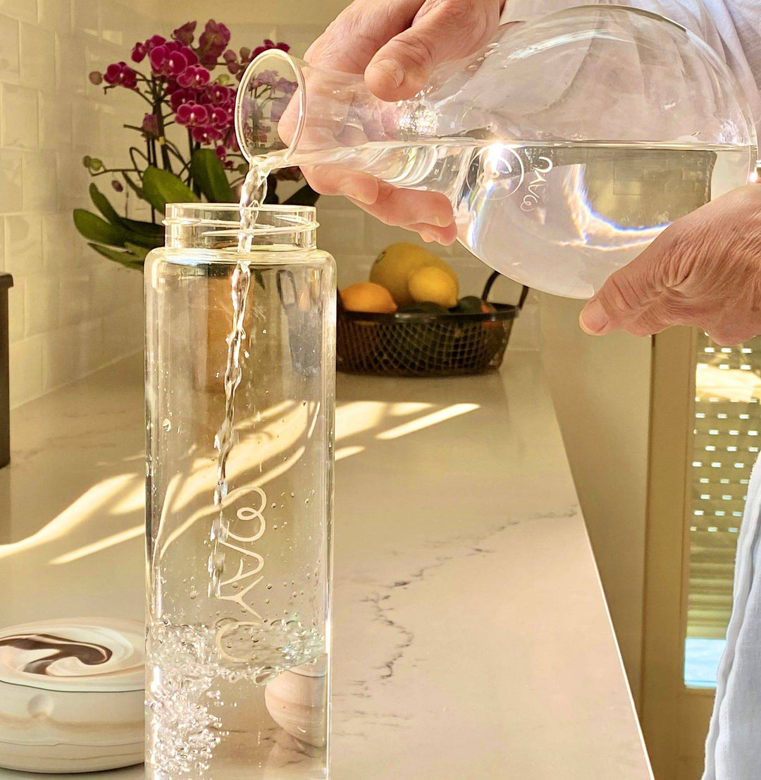 Someone pours water from a glass pitcher into the Mayu Water Pure Glass Bottle, a tall, BPA-free bottle, on the kitchen counter. In the background, lemons in a fruit basket and purple orchids are visible as sunlight streams in.