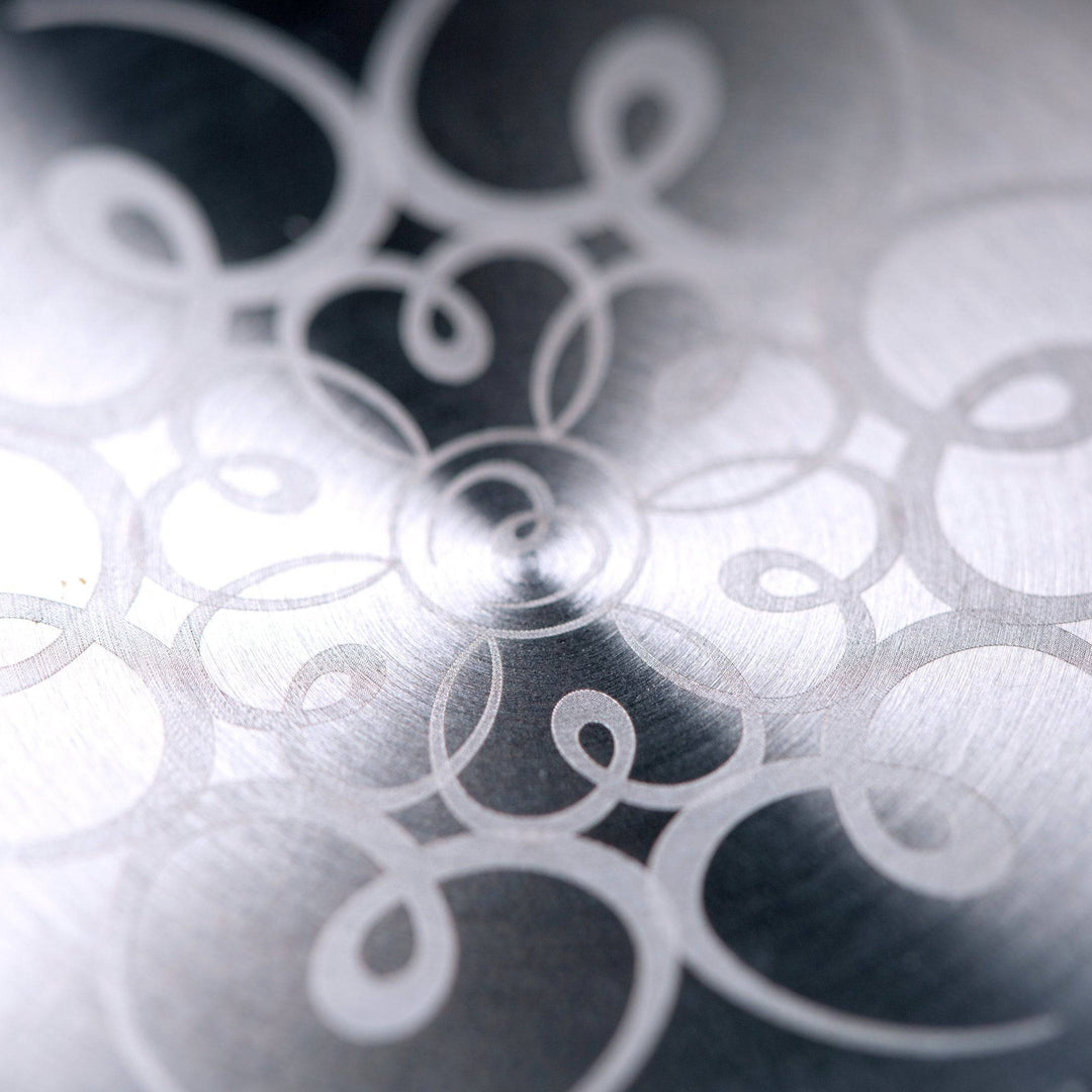 Close-up of an intricate, swirling pattern etched onto brushed metal, echoing the elegant, curving lines found on Mayu Water’s Pure Glass Bottle.