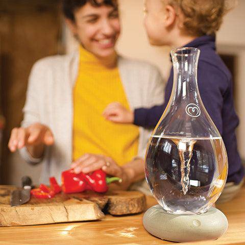 A Mayu Water Swirl Glass Carafe with its signature vortex sits on a table, while in the softly blurred background, a smiling adult and child prepare food together in a cozy kitchen.