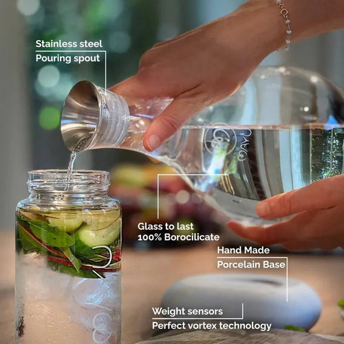A hand pours water from the Mayu Water Swirl Structured Water Carafe, featuring a handblown borosilicate glass body, stainless steel spout, porcelain base, and weight sensors—all designed to enhance hydration quality.
