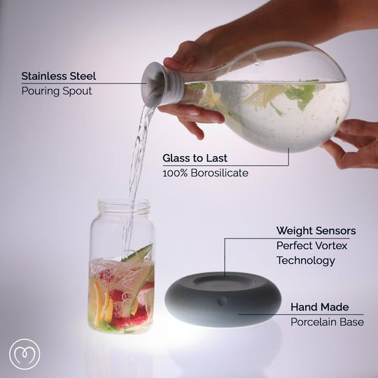 A hand pours water from the MAYU Swirl Carafe | Onyx by Mayu Water with a stainless steel spout into a fruit-filled jar, creating balanced hydration. The round gray porcelain base showcases weight sensors, borosilicate glass, and handmade porcelain.