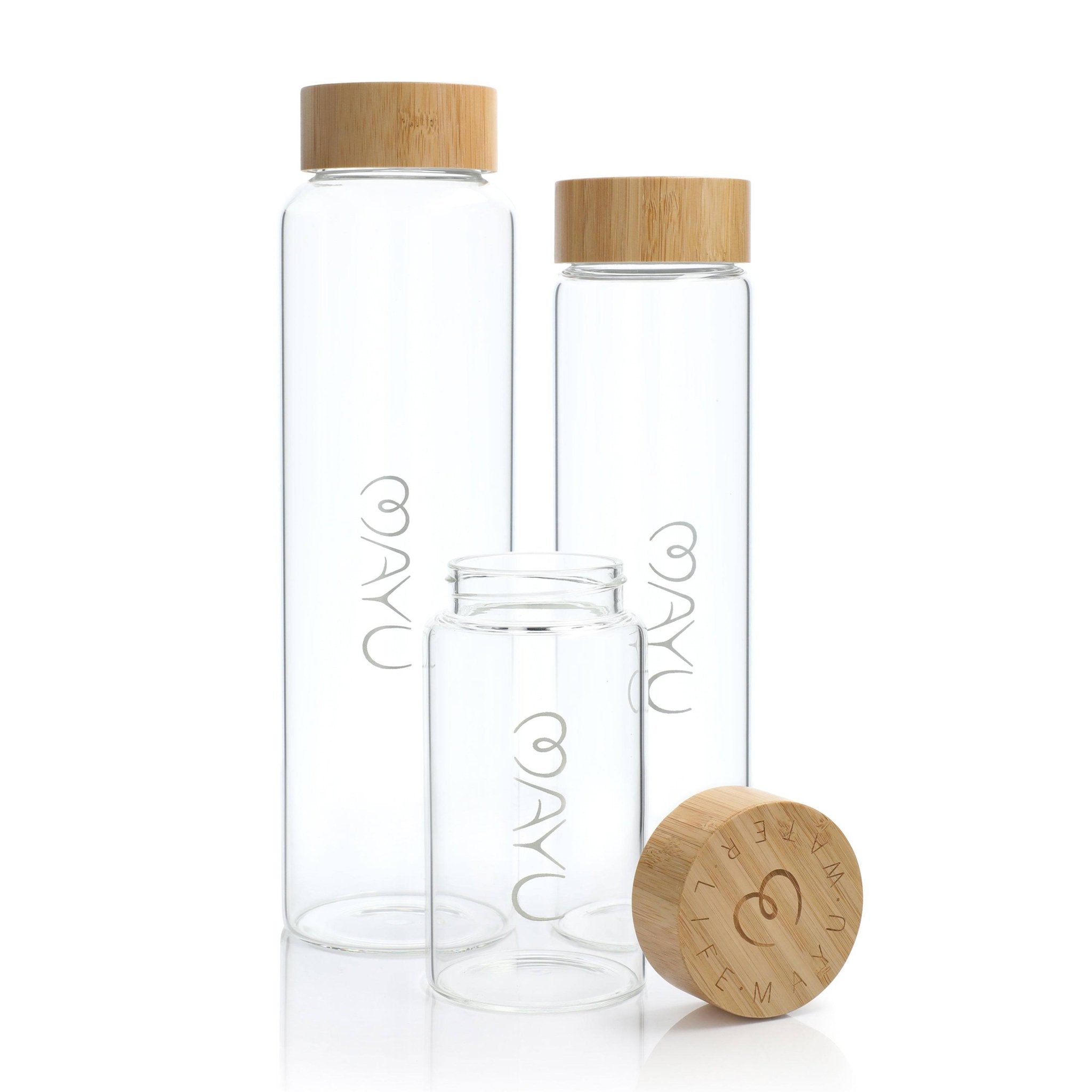 Three Pure Glass Bottles by Mayu Water with light wooden lids: two stand upright and one lies sideways, its BPA-free lid removed to display a circular wood top engraved with intricate markings.