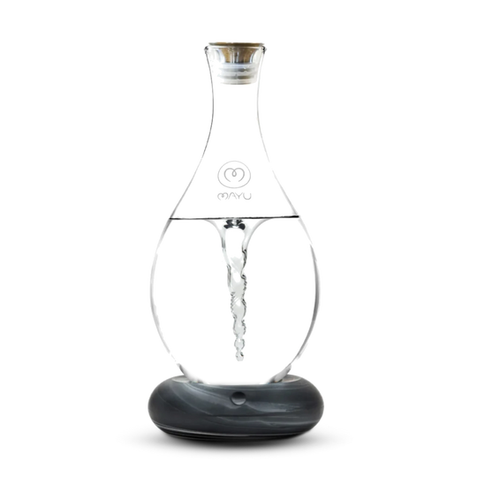 The Mayu Water Swirl Structured Water Carafe, with a lid and round dark base, creates a visible water vortex inside its clear glass body. The product is shown against a plain white background.