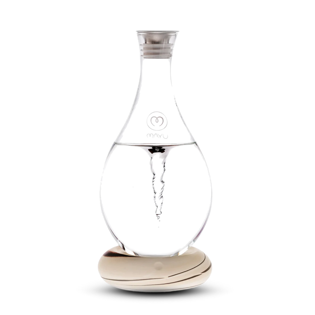 The Swirl Structured Water Carafe by Mayu Water features a clear glass body with a swirling vortex inside, silver cap, and Mayu logo. It sits on a patterned light wood base against a white background.