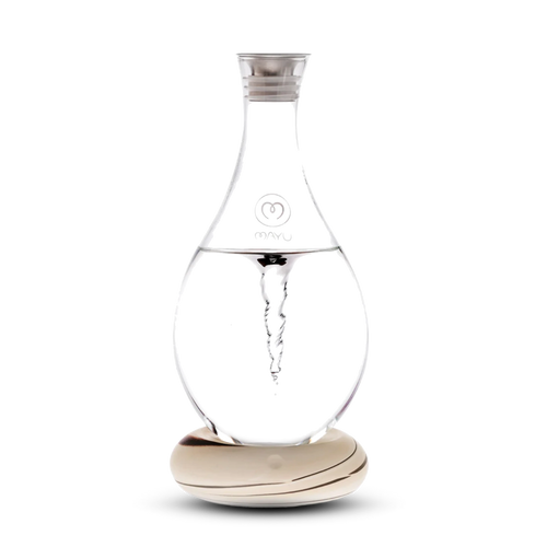 The Swirl Structured Water Carafe by Mayu Water features a clear glass body with a swirling vortex inside, silver cap, and Mayu logo. It sits on a patterned light wood base against a white background.