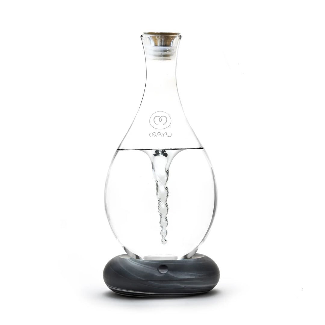The MAYU Swirl Carafe by Mayu Water features a clear glass design with a vortex effect, a narrow neck, and a small front logo. It sits on a round gray and white marbled base to enhance your drinking water experience.