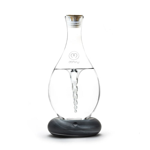 The MAYU Swirl Carafe by Mayu Water features a clear glass design with a vortex effect, a narrow neck, and a small front logo. It sits on a round gray and white marbled base to enhance your drinking water experience.