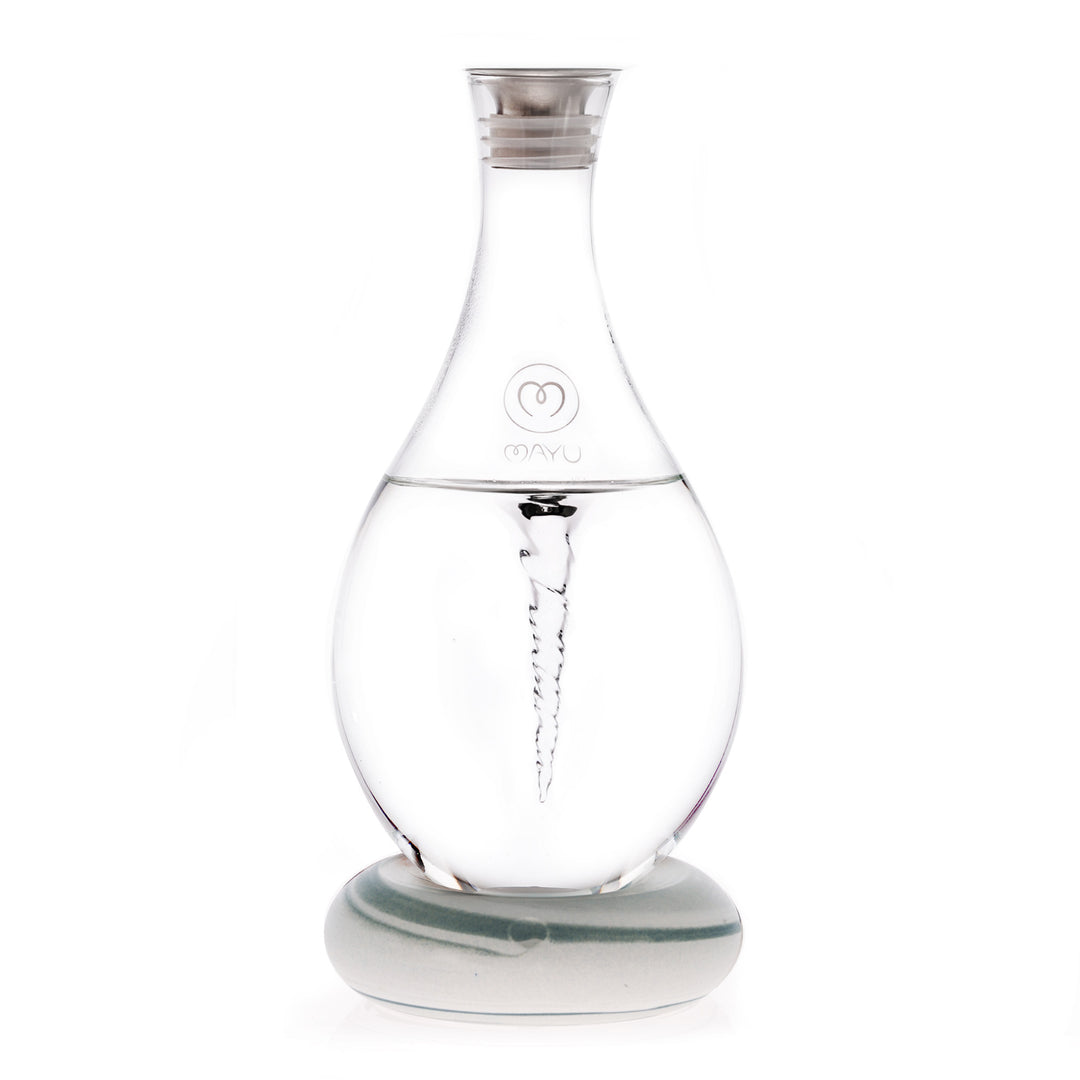 The MAYU Swirl Carafe by Mayu Water is a clear glass carafe with a silver cap on a round base. It creates a visible water vortex to enhance drinking water and features the MAYU logo on its front.