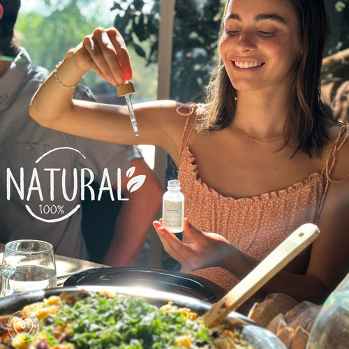 A smiling woman adds Mayu Water Essential Minerals - Single to her meal from a dropper bottle at a sunlit table. “Natural 100%” is written on the left. In the foreground are a glass of water and a dish garnished with greens.