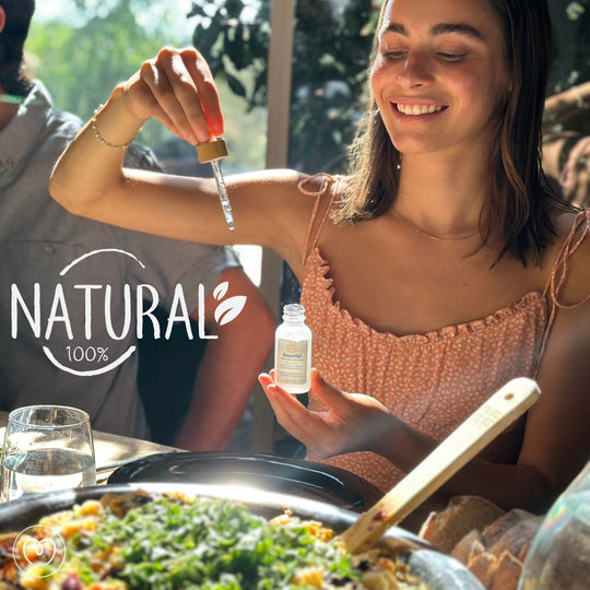 A smiling woman adds Mayu Water Essential Minerals - Single to her meal from a dropper bottle at a sunlit table. “Natural 100%” is written on the left. In the foreground are a glass of water and a dish garnished with greens.