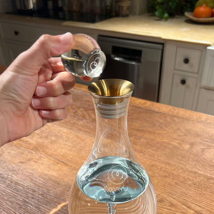 A hand pours water from the Mayu Water Glass Topper with a stainless steel spout into a swirl carafe on a wooden kitchen counter, with white cabinets, a dishwasher, and a fruit bowl in the background.