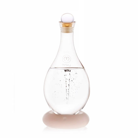 The Mayu Water Glass Topper, a clear teardrop-shaped swirl water carafe with a limited edition glass topper and a small vortex inside, sits on a smooth round white base against a plain white background.