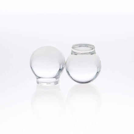 Two small, round, transparent Glass Topper jars by Mayu Water sit side by side on a white surface—one upright, one upside down—reflecting light with elegance.
