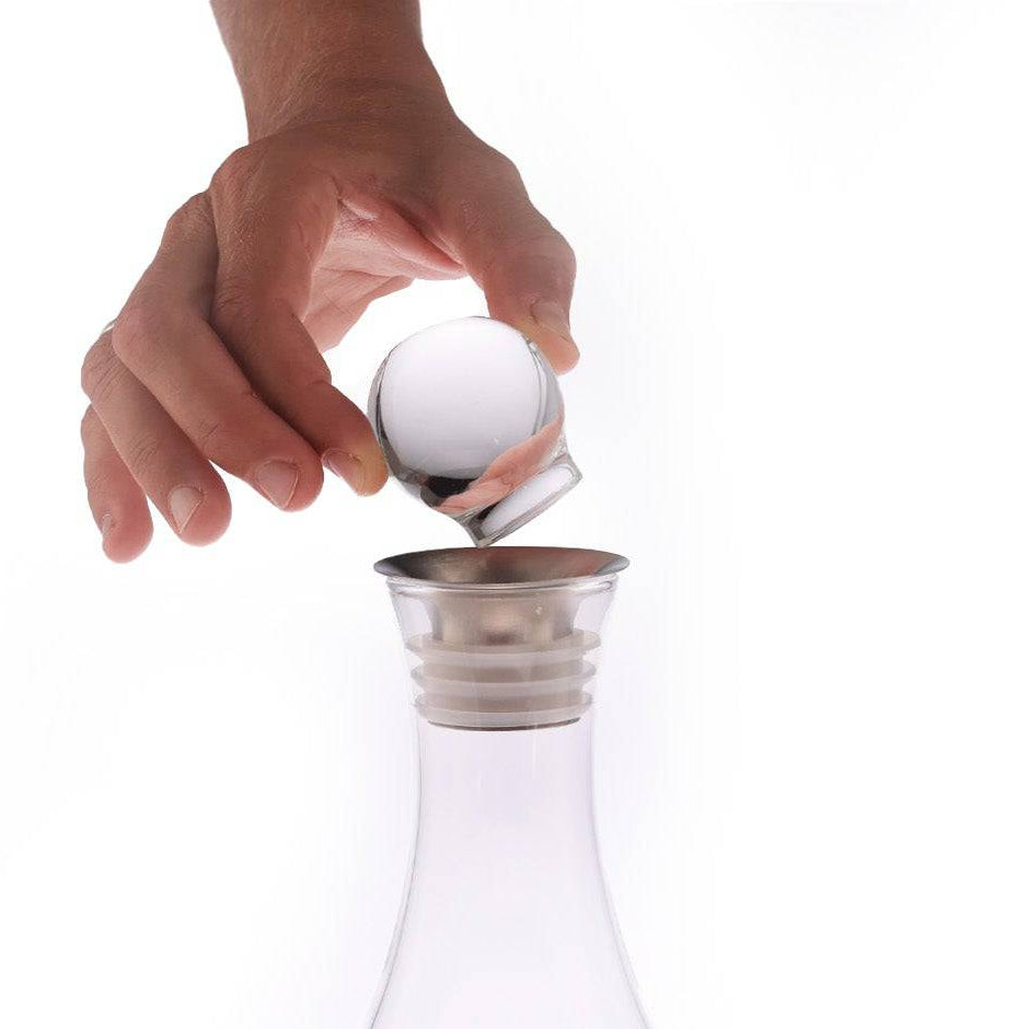 A hand lifts the Mayu Water Glass Topper from the Swirl Carafe, which sits on its round base. The carafe, filled with swirling water that forms a vortex, stands out against a white background.