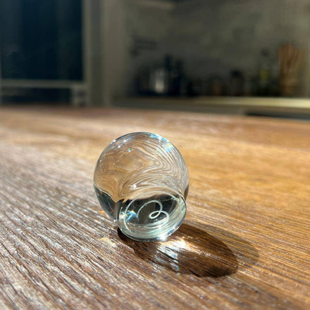 A clear glass marble with swirling patterns rests on wood, reflecting light like the Mayu Water Glass Topper. A blurred kitchen background adds depth to this tranquil scene.