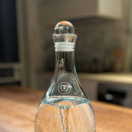 A Mayu Water Glass Topper filled with water rests on a wooden kitchen surface. The clear carafe has a round glass stopper and features a white heart logo on its neck, with blurred kitchen items visible in the background.