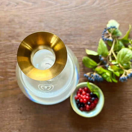 A top-down view of Mayu Water's Swirl Golden Pouring Spout on a glass bottle, alongside a bowl of red and black berries and green leaves with blue berries, all beautifully arranged on a wooden surface.