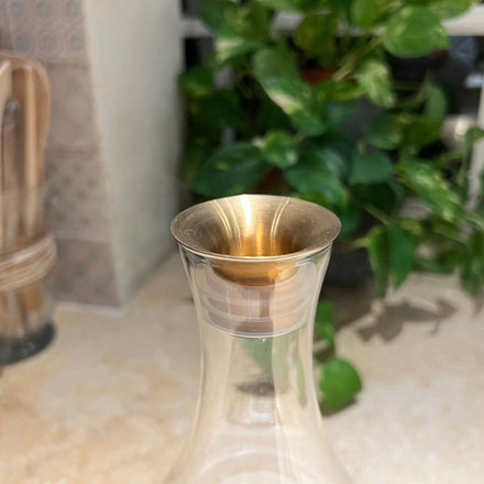 A close-up of the Mayu Water carafe fitted with the Swirl Golden Pouring Spout rests on a beige countertop, with a leafy green plant and kitchen utensils gently blurred in the background.