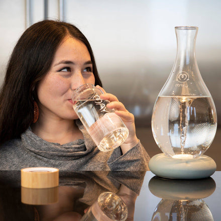 A woman with long dark hair enjoys water from a Mayu Water Pure Glass Bottle, sitting beside a decorative carafe at a bright, modern table. The glass bottle is eco-friendly and BPA free.