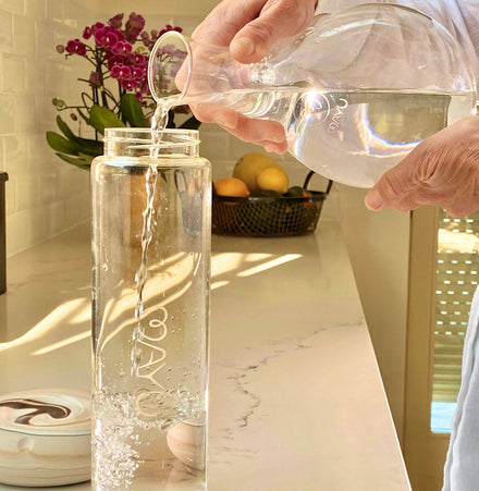 Someone pours water from a glass pitcher into the Mayu Water Pure Glass Bottle, a tall, BPA-free bottle, on the kitchen counter. In the background, lemons in a fruit basket and purple orchids are visible as sunlight streams in.