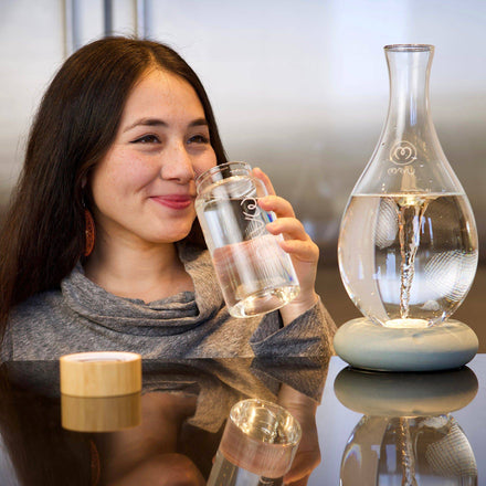 A woman with long dark hair smiles while holding a glass of water. Nearby on the table, a Mayu Water Swirl Glass Carafe made of borosilicate glass rests next to its wooden lid in a bright, modern setting.