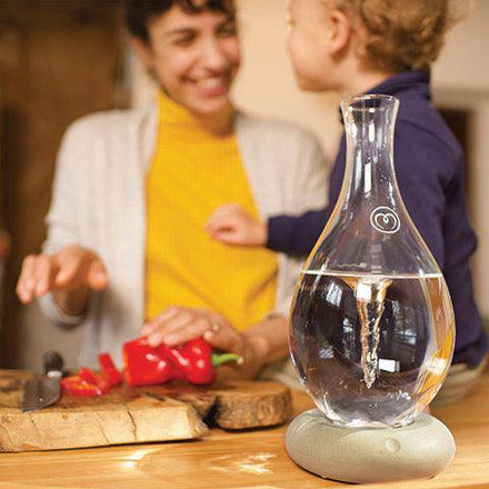 A Mayu Water Swirl Glass Carafe with its signature vortex sits on a table, while in the softly blurred background, a smiling adult and child prepare food together in a cozy kitchen.