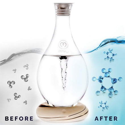 The Mayu Water Swirl Structured Water Carafe, made of handblown borosilicate glass with a swirling vortex and wooden base, is shown with gray molecules (before) and blue molecules (after) to illustrate improved drinking water quality. Labels BELOW read BEFORE and AFTER.