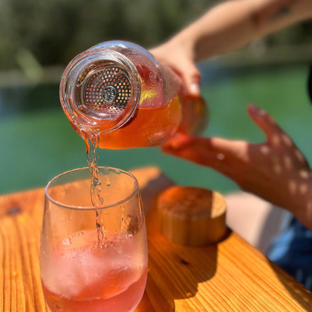 Someone pours a pink drink from a Mayu Water Pure Glass 1.5L Bottle (Pack of 3) into a tumbler with ice on a wooden table outdoors, with a blurred green background.