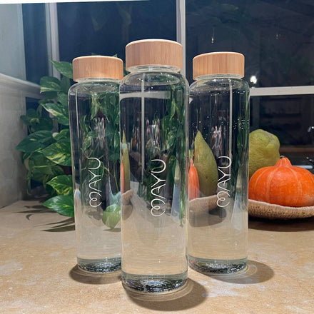 Three Mayu Water Spin the Bottles glass bottles with wooden lids sit on a kitchen counter, reflecting an eco-conscious lifestyle. Green plants, a pear, and an orange pumpkin decorate the background beneath a window.