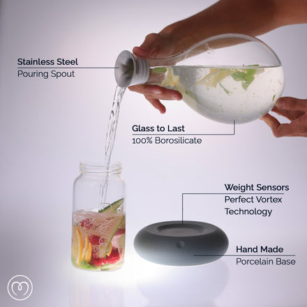 A hand pours water from the MAYU Swirl Carafe | Onyx by Mayu Water with a stainless steel spout into a fruit-filled jar, creating balanced hydration. The round gray porcelain base showcases weight sensors, borosilicate glass, and handmade porcelain.