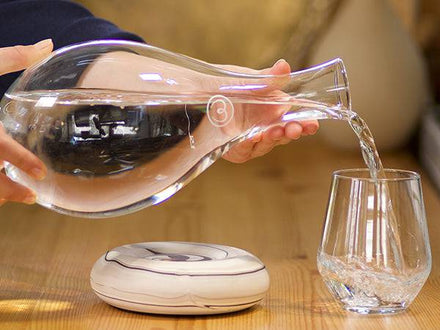 A person pours water from a Mayu Water Swirl Glass Carafe into a glass on a wooden table, with a round ceramic object nearby.